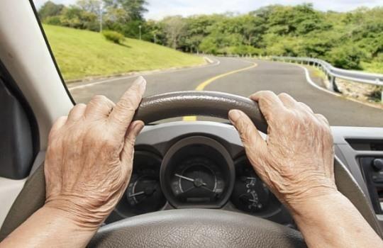 一位老人正開車行駛在一條蜿蜒的鄉間道路上，畫面展現了韓國高齡駕駛的現象。